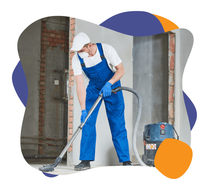 a man in blue overalls wearing a mask and vacuuming post renovation dust off of a floor.